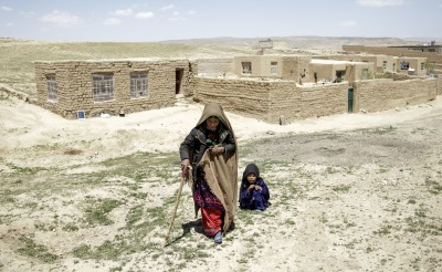 A mother walks away from her home with the help of a cane. Her daughter crouches behind her.