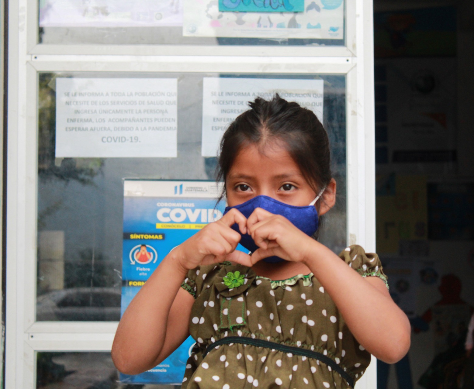 A young girl, wearing a mask, makes a heart with her hands.