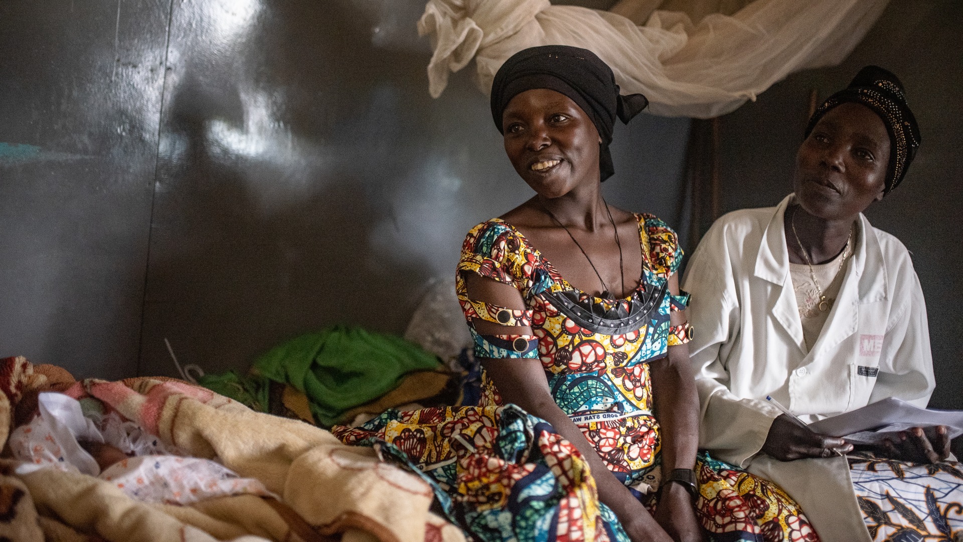 A woman who recently gave birth sits on a bed with her nurse and baby.