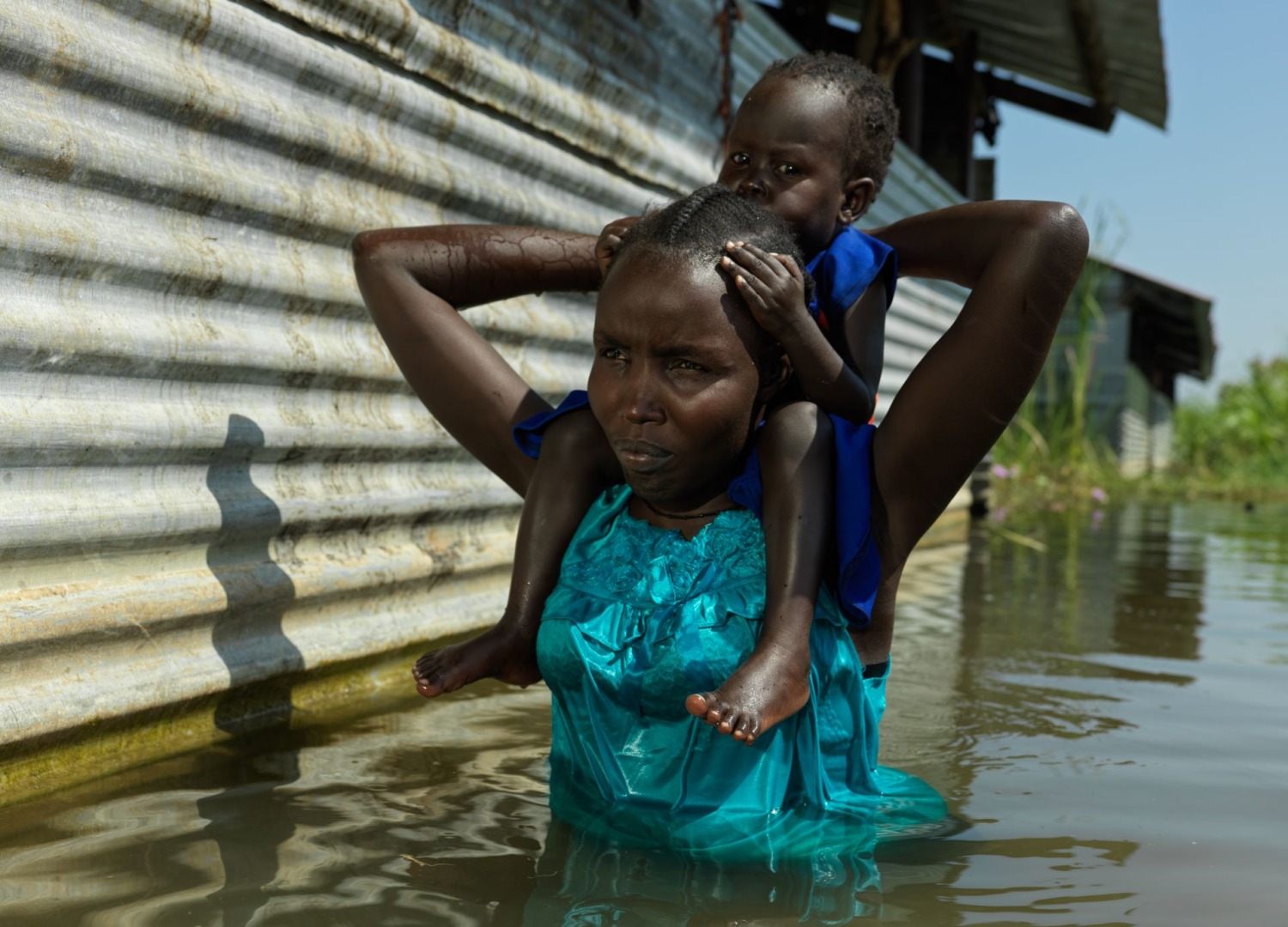 A woman holding a child on her shoulders, walking through the waist-high flood in South Sudan.