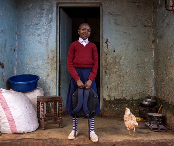 A girl in Tanzania stands ourside her front door holding a backpack