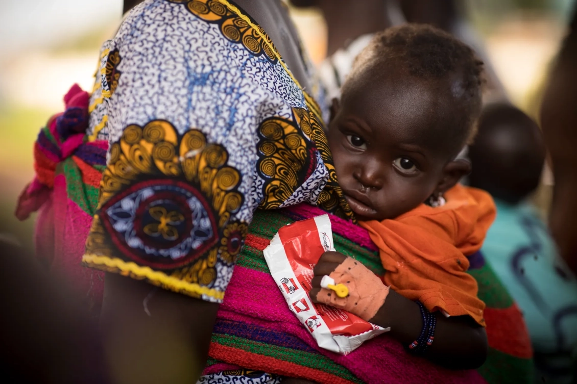 A child, carried by his mother, holds a packet of nutrition treatment